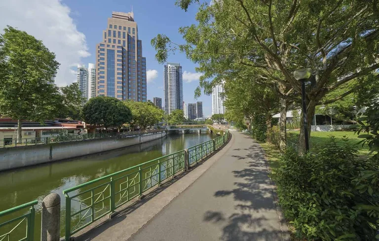 Photograph of the area, for illustration only: a shaded path of the park connector along the canal, with trees on one side and towers beyond